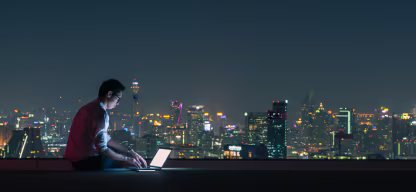 A person using a laptop on a rooftop at night with a city skyline in the background
